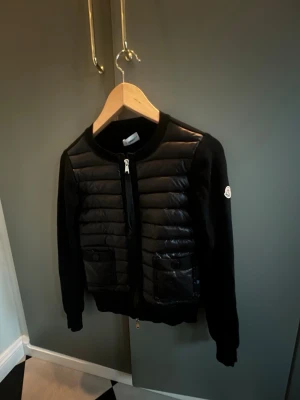 Svart cardiganjacka från Moncler - Svart cardiganjacka från Moncler med quiltade partier framtill och två fickor med knappstängning. Jackan har rund halsringning, dragkedja och Moncler-logga på ärmen. Andvänd fåtal gånger och i ny skick.jättesnygg till vår/sommar och passar med allt skriv för bilder❤️