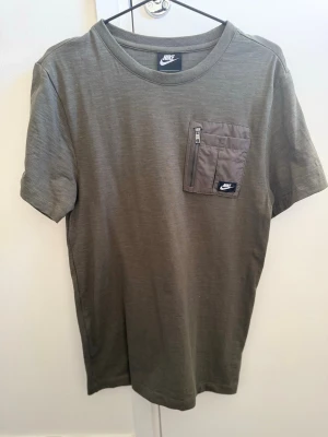 Olivgrön Nike t-shirt med ficka XS - Olivgrön t-shirt från Nike med rund halsringning och korta ärmar. Bröstficka med dragkedja och Nike-logga ger en sportig touch. Tillverkad i mjukt material som känns skönt mot huden. Perfekt för en avslappnad och trendig stil.