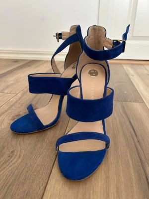 Blåa pumps från River Island - Snygga blåa pumps från River Island i mjuk mocka med tre breda remmar och smal klack. Skorna har öppen tå och justerbart spänne runt ankeln för extra support. Perfekta för dig som vill sticka ut med färg och stil. Storlek 5. 
