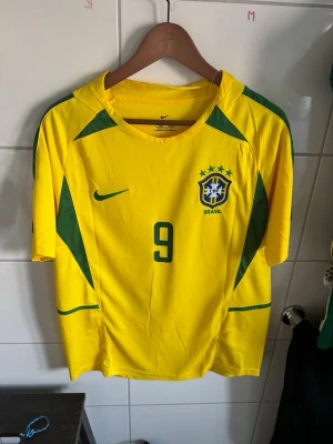 Brasilien Ronaldo 9 matchtröja Nike - Gul Brasilien fotbollströja från Nike med gröna detaljer, nummer 9 och Ronaldo på ryggen. Klassisk landslagströja med CBF-emblem och korta ärmar. Perfekt för dig som älskar fotboll och vill sticka ut på planen eller läktaren.
