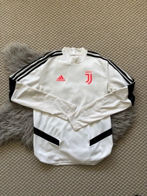 Vit Juventus träningströja Adidas S - Vit långärmad träningströja från Adidas med Juventus-logga och Adidas-märke i rosa på bröstet. Svarta ränder längs ärmarna och svarta detaljer vid fickorna. Tillverkad i lätt och funktionellt material, perfekt för träning eller fotboll.