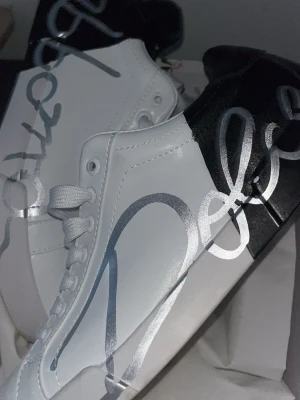 Dolce & Gabbana vita sneakers - Snygga vita sneakers från Dolce & Gabbana med svart häl och stora silvriga logotyp-prints över sidorna. Skorna har klassisk låg modell, vita snören och är tillverkade i slätt läder. Perfekta för dig som vill sticka ut med en exklusiv touch.