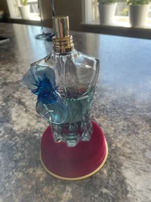 Jpg Le Beau Flower Edition - Unik parfymflaska formad som en manlig torso i genomskinligt glas med blågrön vätska och en blå blomdekoration. Kommer i en snygg cylinderformad förpackning med blommotiv och gulddetaljer. Perfekt för dig som gillar exklusiva och kreativa dofter.