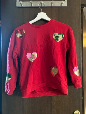 Röd sweatshirt med paljett-hjärtan - Cool röd sweatshirt med flera hjärtformade paljetter i guld, rosa och grönt på framsidan och ärmarna. Tröjan har rund hals och ribbade muddar. Perfekt för dig som gillar färg och glitter! Materialet är mjukt och bekvämt.