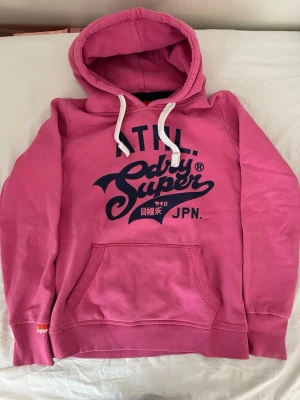 Rosa hoodie från Superdry med tryck - Cool rosa hoodie från Superdry med stort mörkblått tryck på bröstet och japanska tecken. Hoodien har vit snörning i huvan och en klassisk känguruficka framtill. Perfekt för dig som gillar streetstyle och vill ha något bekvämt och snyggt.