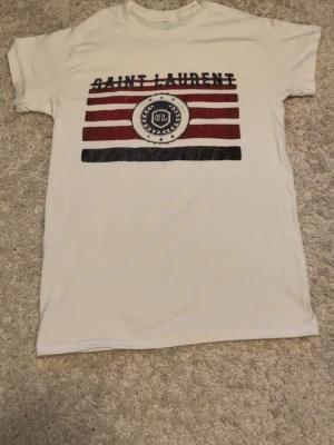 Vit Saint Laurent t-shirt med tryck - Vit t-shirt från Saint Laurent med tryck i rött, svart och vitt framtill. T-shirten har rund halsringning och korta ärmar. Trycket består av ränder och en cirkel med SL-logga. Perfekt för en cool och avslappnad stil. Är lite osäker på äkthet därför säljer jag billigt, bara att hojta till för fler frågor