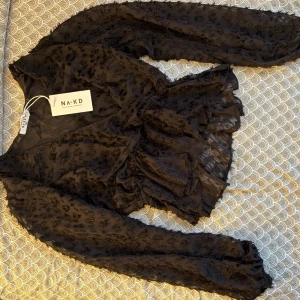 Svart omlottblus med volang från NA-KD ( NY MED PRISLAPP) - Snygg svart omlottblus från NA-KD med transparent tyg och diskret blommigt mönster. Blusen har långa ärmar, volangdetaljer och knyt i midjan. Perfekt för dig som vill ha en trendig och feminin look.