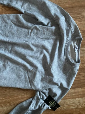 Grå sweatshirt från Stone Island - Snygg grå sweatshirt från Stone Island med klassisk rund halsringning och ikonisk logotyp-patch på ärmen. Tillverkad i mjukt bomullsmaterial som ger en avslappnad och stilren look. Perfekt till jeans eller joggers.