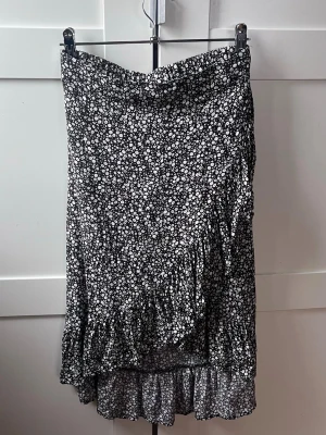 Svart blommig långkjol med volang - Svart långkjol med vitt blommönster och volangkant. Kjolen har omlottdetalj framtill och resår i midjan för en bekväm och snygg passform. Tillverkad i ett lätt och luftigt material som känns perfekt för sommaren. Kjolen är i stl 146/152, alltså barnstorlek och då är den ungefär till anklarna. Men den går även att använda om du har en större storlek än så då det är resår i midjan och snygg även om den blir lite kortare