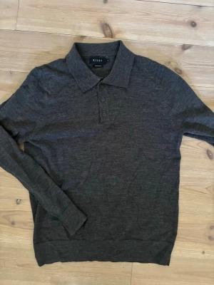 Mörkgrå stickad tröja från Riley - Mörkgrå stickad tröja från Riley i mjuk merinoull. Tröjan har klassisk krage och knäppning med tre knappar framtill. Långärmad modell med ribbade muddar vid ärmslut och nederkant. Perfekt för en stilren och bekväm look.