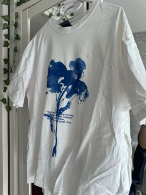 Vit t-shirt - Vit t-shirt med ett stort blått blommotiv och text på framsidan. T-shirten har rund hals och korta ärmar, perfekt för en avslappnad stil. Tillverkad i mjuk bomull för skön känsla hela dagen.