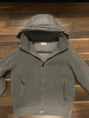 Grå pufferjacka från Moncler - Snygg grå pufferjacka från Moncler med stickade ärmar och ribbade muddar. Jackan har vadderad kropp och huva, två framfickor med dragkedja och en hög krage. Perfekt för kalla vinterdagar och riktigt trendig look.