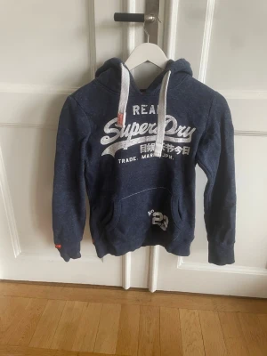 Blå Superdry hoodie med tryck - Mörkblå hoodie från Superdry med vit logga och text på bröstet samt vita dragsnören i huvan. Klassisk känguruficka framtill och ribbade muddar vid ärmar och nederkant. Perfekt för en avslappnad och sportig stil.