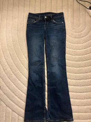 Bootcut jeans med broderade bakfickor - Mörkblå bootcut jeans med snygga slitningar och broderade detaljer på bakfickorna. Klassisk femficksmodell med låg midja och dragkedja. Perfekta för en avslappnad och trendig look.