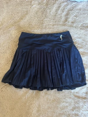 Svart plisserad sportkjol RS Sports - Svart sportkjol från RS Sports i storlek S. Kjolen är plisserad och har bred resår i midjan för extra komfort. Tillverkad i ett lätt och stretchigt material som passar perfekt för tennis eller padel. Diskret logga framtill. Inbyggda shorts.