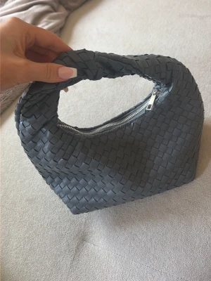 Grå flätad handväska med dragkedja - Snygg grå handväska i flätad design med ett mjukt, böjt handtag och silverfärgad dragkedja. Väskan har en modern och trendig look, perfekt för att lyfta din outfit. Materialet är konstläder och väskan har en kompakt, stilren form.