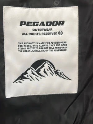 Svart vindjacka från Pegador - Säljer en svart vindjacka från Pegador med coolt innerfoder och dragkedja. Jackan är tillverkad i polyester och har en urban look, perfekt för dig som gillar äventyr och streetwear. Stora loggor på insidan och snygga detaljer.