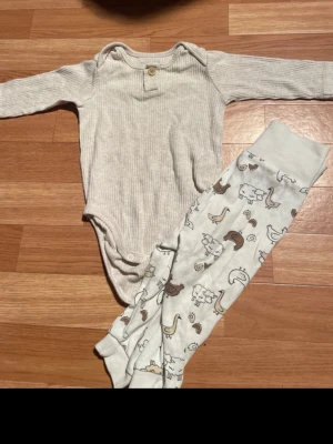 Randig body och byxa med djurmönster - Säljer ett set med en ljusgrå, ribbad body med lång ärm och knappdetalj vid halsen samt ett par vita byxor med sött djurmönster i brunt och beige. Perfekt för mysiga dagar hemma. Materialet känns mjukt och bekvämt mot huden.
