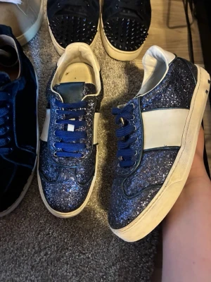 Valentino flycrews  - Snygga sneakers med blå glitteryta, vita paneler på sidorna och blå snörning. Skorna har en klassisk låg profil och vit sula, perfekta för att sticka ut i mängden. Materialet är syntet med glittereffekt och textilfoder. Box medföljer vid köp 