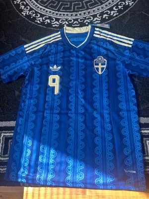 Blå Sverige fotbollströja Adidas - Blå fotbollströja från Adidas med Sveriges landslagsemblem och nummer 9 på bröstet med namnet Isak på ryggen samt nummer 9. Tröjan har vita detaljer, klassiska Adidas-ränder på axlarna och ett retroinspirerat mönster. Tillverkad i lätt och ventilerande ClimaCool-material.