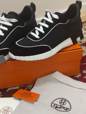 H Skor  - Snygga svarta sneakers  med vit sula och vita detaljer. Skorna har vita snören och en unik sula med H-mönster undertill. Materialet är en mix av textil och läder, och designen är modern med kontrasterande linjer.
