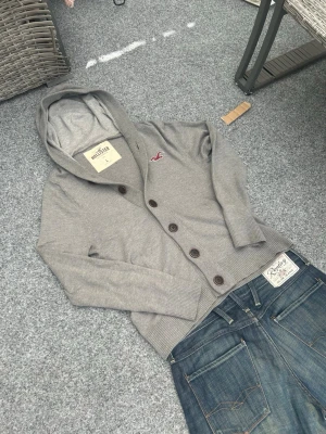 Hollister grå cardigan hoodie L - Grå cardigan/hoodie från Hollister i mycket bra skick. Stilren modell med knappar framtill istället för dragkedja, vilket ger en mer clean och preppy look. Har även huva och broderad logo på bröstet.  Storlek L men funkar även som en lite oversized fit på M. Perfekt för layering och passar till både jeans och mer uppklädda outfits.  Mjuk och bekväm – perfekt för vardag, streetwear eller casual stil.  Skick: mycket bra