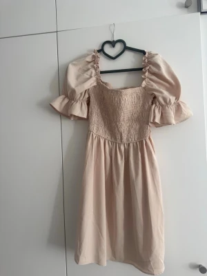 Ljusbeige klänning med puffärmar - Supersöt ljusbeige klänning med smockat liv och voluminösa puffärmar. Klänningen har fyrkantig ringning och volangdetaljer vid ärmsluten. Perfekt för sommarens alla tillfällen!