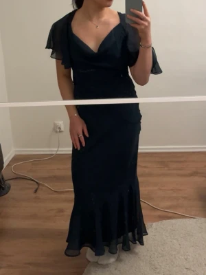 Mörkgrön långklänning med öppen rygg - Långklänning med v-ringning och volangärmar. Klänningen har öppen rygg med  snörningsdetaljer och ger en fin siluett. Har aldrig använts, även prislappen är kvar. 