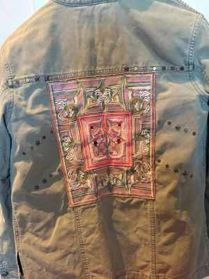 Olivgrön oddmolly jeansjacka med brodyr - Unik olivgrön jeansjacka med stor färgglad broderad patch på ryggen och små nitar i stjärnform. Jackan har klassisk krage, knappstängning framtill och dekorativ patch även på ena ärmen. Perfekt för dig som vill sticka ut med coola detaljer.