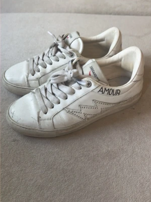 Vita sneakers Zadig & Voltaire, köpte för 2800kr - Säljer ett par vita sneakers från Zadig & Voltaire med svart och röd ZV-logga på hälen, texten 'AMOUR' på sidan och blixtdetalj. Skorna har vita snören och är tillverkade i läder. Perfekta för dig som gillar en clean men ändå unik look.