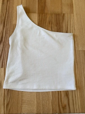 Vit ribbad oneshoulder topp - Snygg vit ribbad topp med oneshoulder-design. Perfekt för dig som vill ha en stilren och trendig look. Toppen är ärmlös och har en smickrande passform som passar till många olika outfits.