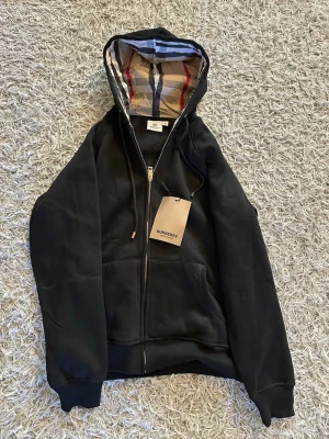 Svart hoodie från Burberry med dragkedja - Svart hoodie från Burberry med klassiskt rutigt foder i huvan och dragkedja framtill. (Står XL men är som S). Mått kan såklart skickas. Pris kan diskuteras. (Strl S)