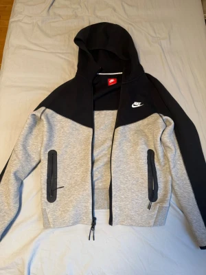 Nike tech fleece  - Snygg hoodie från Nike i grått och svart med dragkedja framtill och två fickor med svarta dragkedjor. Klassisk Nike-logga broderad på bröstet. Hoodien har en stor huva och är gjord i mjukt sweatshirtmaterial, perfekt för chill eller träning. Haft den på mig 2-3 gånger annars helt ny, köpt från Nikes hemsida alltså äkta.
