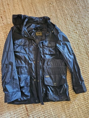 Polo Ralph Lauren vindjacka stl 10-12 - Snygg mörkblå vindjacka från Polo Ralph Lauren med fyra stora fickor framtill, dragkedja och knappar. Jackan har huva och justerbar midja för skön passform. Tillverkad i lätt, glansigt material som står emot vind. Perfekt för dig som vill ha en streetig och funktionell look.