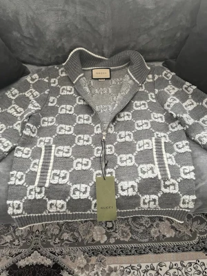 Gucci tröja  - Snygg grå stickad kofta från Gucci med vit GG-logga över hela plagget. Koftan har hög krage, ribbade muddar och dragkedja framtill. Två fickor på sidorna och exklusiv känsla. Perfekt för dig som vill ha något extra från ett lyxigt märke. Helt ny och tag finns kvar!