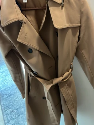 Klassisk beige trenchcoat med bälte - Stilren beige trenchcoat med dubbelknäppning och matchande bälte i midjan. Jackan har bred krage, svarta knappar och raka ärmar. Perfekt för dig som vill ha en tidlös och trendig look. Materialet känns mjukt och följsamt. Andvänd sparsamt 