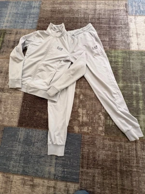 Grått EA7 mjukisset med dragkedja - Snyggt grått mjukisset från EA7 med jacka som har dragkedja och ståkrage samt matchande byxor med text på ryggen . Båda delarna har EA7-logga på bröstet och benet. Setet är gjort i mjukt och stretchigt material som är skönt att ha på sig.