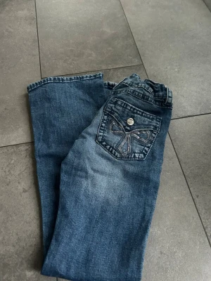 Blå jeans med broderade bakfickor från Gina tricot - Säljer ett par blå jeans med snygga broderade detaljer på bakfickorna och silvriga knappar. Jeansen har en klassisk rak passform och är tillverkade i denim. Perfekta för dig som vill ha ett par stilrena men ändå unika jeans i garderoben.