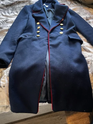 Marinblå rock med guldknappar sopran - Stilren marinblå rock med dubbelknäppning och sex guldknappar framtill. Rock sopran med röd piping längs krage och slag, samt dekorativa guld pipeline sum knappar på ärmen. Fodrad insida och klassisk militärinspirerad design. Perfekt för dig som vill sticka ut.