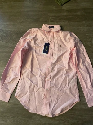 Rosa skjorta från Polo Ralph Lauren - Klassisk ljusrosa skjorta från Polo Ralph Lauren med broderad logga på bröstet. Skjortan har lång ärm, knappar framtill och en tidlös krage. Perfekt för en stilren look och tillverkad i mjuk bomull. Den är skrynklig då jag har ej strykt den eller anvödna på oerhört länge 