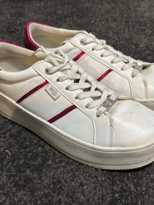 Vita sneakers med rosa detaljer - Snygga vita sneakers från Pernilla Wahlgren med rosa metallic-detaljer på sidan och runt hälen. Skorna har klassisk låg profil, vita snören och en tjock sula. Tillverkade i konstläder med diskret logga på sidan. Perfekta för en clean lock.