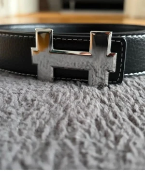 Svart skärp med silverfärgat H-spänne - Stilrent svart skärp med ett ikoniskt H-format spänne i silverfärg. Skärpet är tillverkat i skinn med vit kontrastsöm längs kanterna. Perfekt accessoar för att lyfta din outfit.