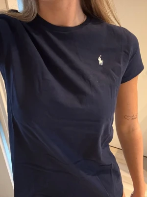 Mörkblå t-shirt från Polo Ralph Lauren - Snygg mörkblå t-shirt från Polo Ralph Lauren med klassisk vit logga broderad på bröstet. T-shirten har rund halsringning och korta ärmar. Tillverkad i mjuk bomull för en skön känsla och perfekt passform. Kom till användning 2 gånger i somras.