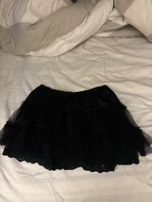 Svart volangkjol i tyll - Säljer en svart kortkjol i tyll med flera lager och volangkant. Kjolen har en fluffig look och passar perfekt för dig som vill ha en edgy och lekfull stil. Elastisk midja för skön passform.