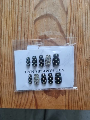 Svarta press-on naglar med pärlor - Snygga press-on naglar i svart med glittriga pärlor och stenar. Några naglar är helt täckta av små rhinestones medan andra har enstaka pärlor för en cool kontrast. Korta till medellånga och i fyrkantig form. Perfekt för dig som vill ha en edgy look.