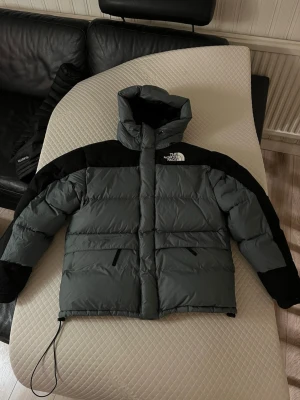 Grå och svart pufferjacka The North Face - Snygg grön/grå pufferjacka från The North Face med svarta detaljer över axlar och ärmslut. Jackan har hög krage, hu Ric, två sidofick niclyckad dragkedja och logga på bröstet autochever. Perfekt för kalla vinterdagar och riktigt streetwear-vibe.