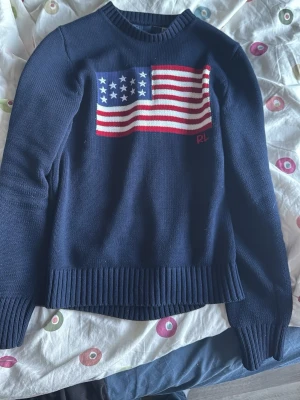 Mörkblå stickad tröja med USA-flagga - Säljer en mörkblå stickad tröja med en stor amerikansk flagga på bröstet. Tröjan har rund hals, långa ärmar och ribbstickade muddar. Perfekt för dig som vill ha en statement-piece i garderoben. Fler frågor finns här 😁