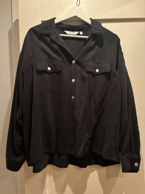 Svart oversized skjorta & Other Stories - Svart oversized skjorta från & Other Stories med klassisk krage, vita knappar och två bröstfickor med lock. Skjortan har lång ärm och avslappnad passform, perfekt för en chill och trendig look. Väldigt skönt material i bomull. 