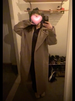 Beige lång ullkappa med bred krage - Stilren beige långkappa med bred krage och stora slag. Kappan har två sidofickor och justerbara band vid ärmsluten. Oversized passform som ger en trendig siluett. Perfekt för kalla dagar och enkel att matcha med olika outfits. Det är i storlek XS Tall 