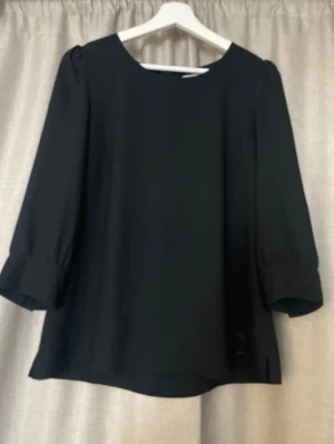  svart blus med trekvartsärm - Stilren svart blus med rund halsringning och trekvartsärmar. Blusen har dekorativa knappar vid ärmsluten och en guldfärgad dragkedja i ryggen. 